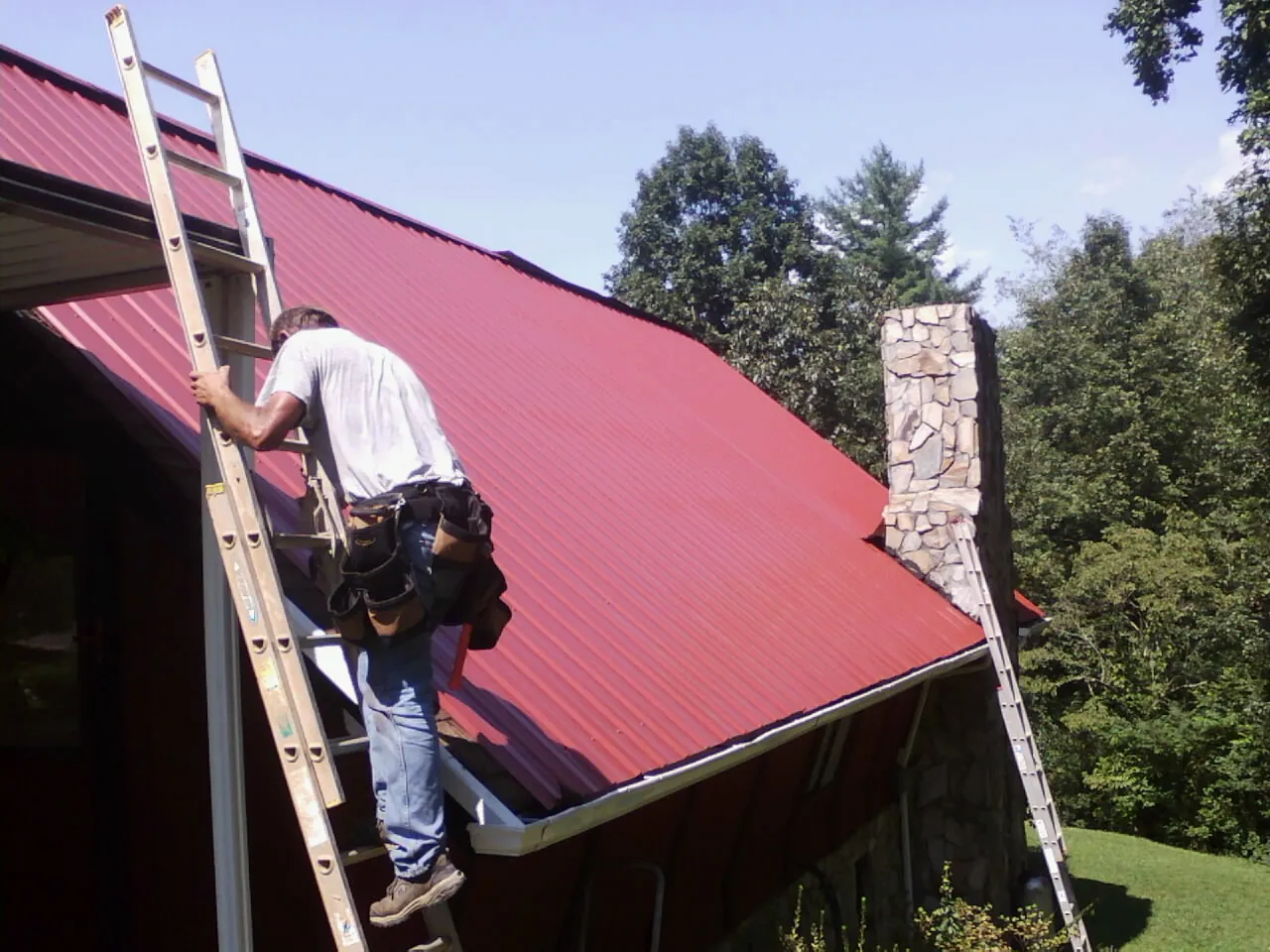 Roofer on a ladder beside a red metal roof and stone chimney — Soffit & Fascia Repair by Roofing Brothers in Glen Burnie