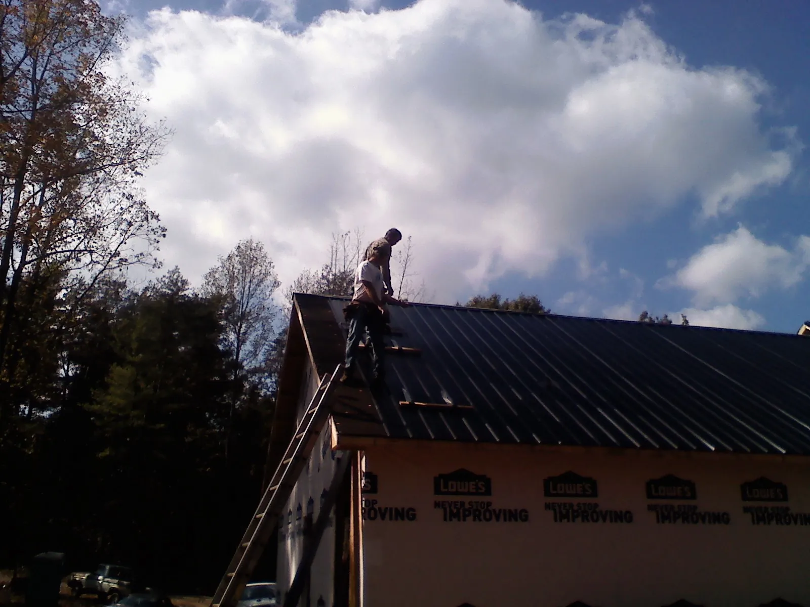 Roofer fastening dark metal panels high against a cloudy sky, from a Roof Inspection job in Glen Burnie